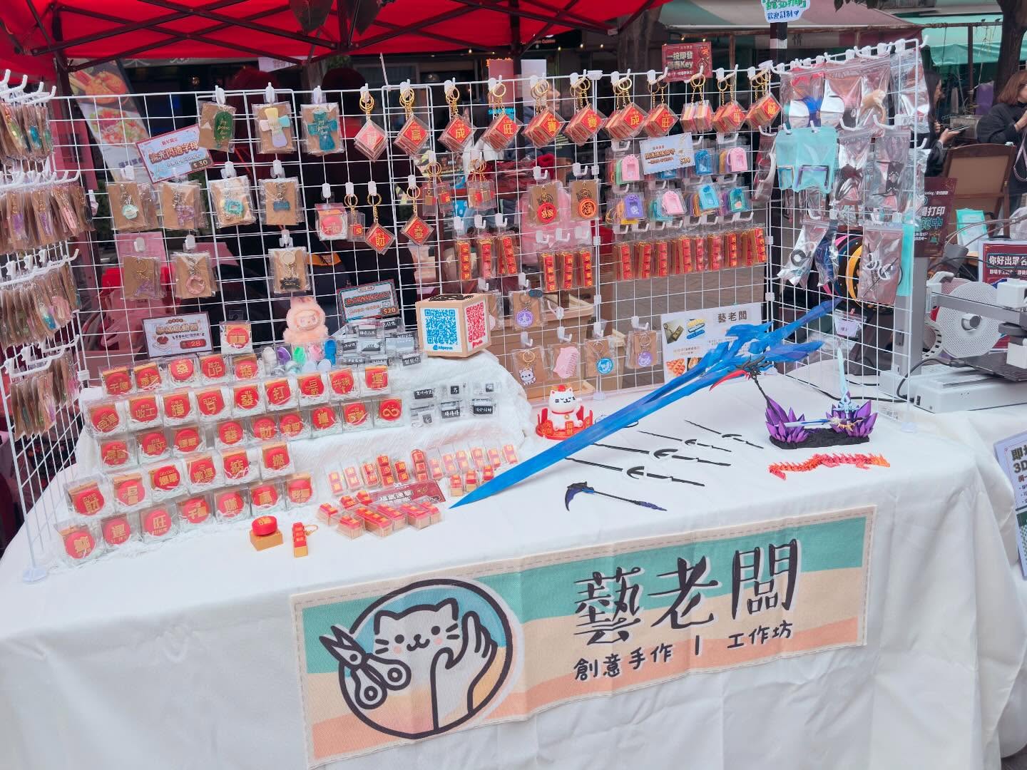 藝老闆市集攤檔：紅色帳篷下的白色網架掛滿手作名牌與匙扣，枱布印有藝老闆貓仔標誌